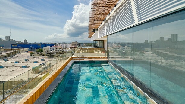 A rooftop swimming pool with a glass barrier, wooden deck, and a view of the cityscape. - 2BR The City Center Dtown Pool Gym Nearcecilip (Santo Domingo)