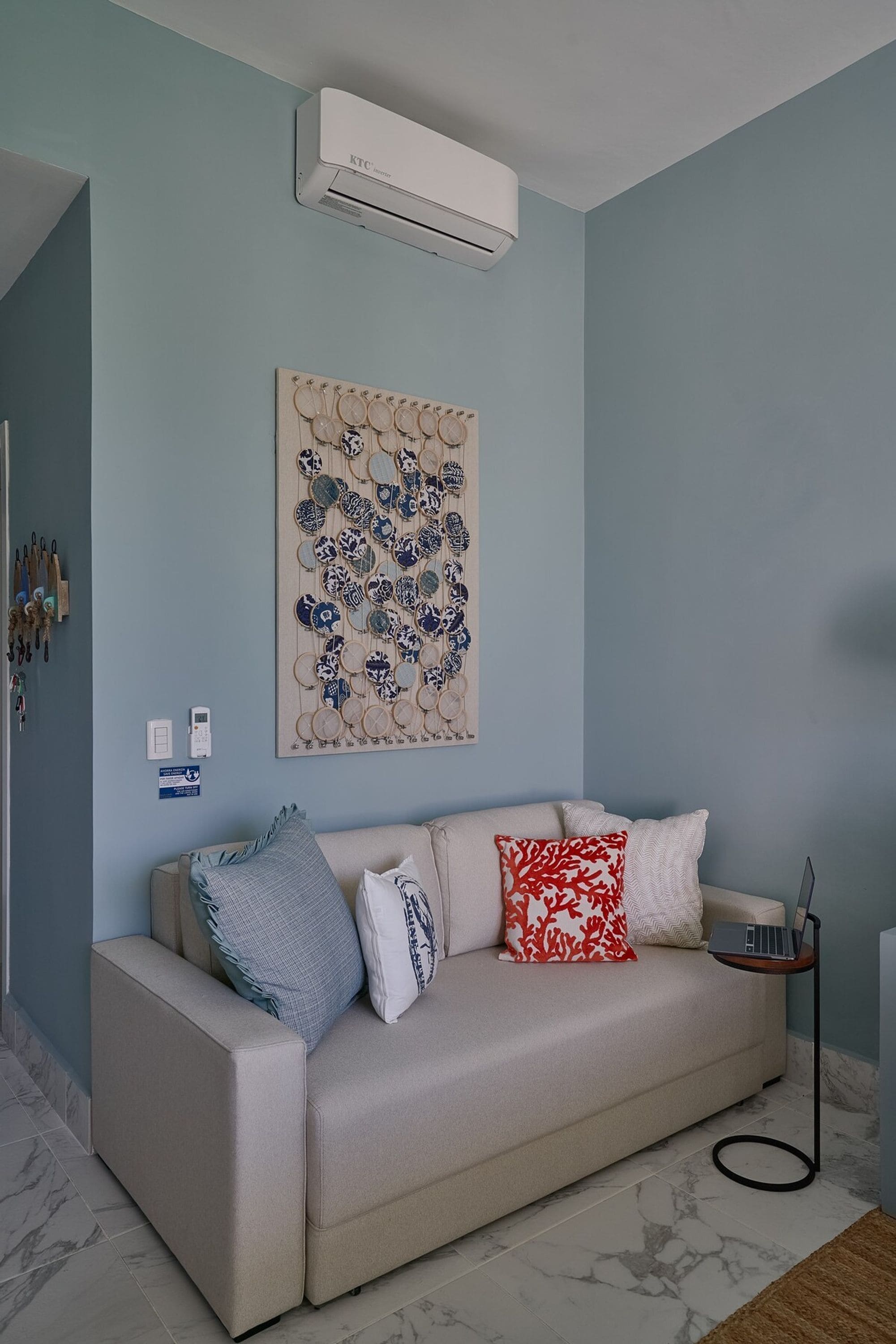 A living room with a beige sofa, a wall-mounted air conditioner, a decorative plate artwork, and a laptop on a stand.