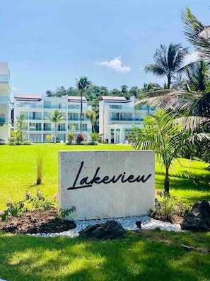 Un letrero con la palabra 'Lakeview' frente a un edificio moderno de varios pisos y grandes ventanales, rodeado de exuberante follaje y palmeras.