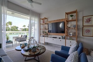 Una sala de estar con televisor, sofá azul y una mesa de centro. Hay estantes con objetos decorativos y una vista a una piscina exterior.