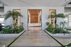 A modern building entrance with a glass door, two palm trees, and a concrete pathway. - Pano View Experience Downtownsd Pool Gym 2BR (Santo Domingo)