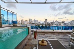 Une piscine sur le toit avec vue sur la ville, un coin salon agrémenté d’un chapeau de paille et un grand verre rempli de boisson.
