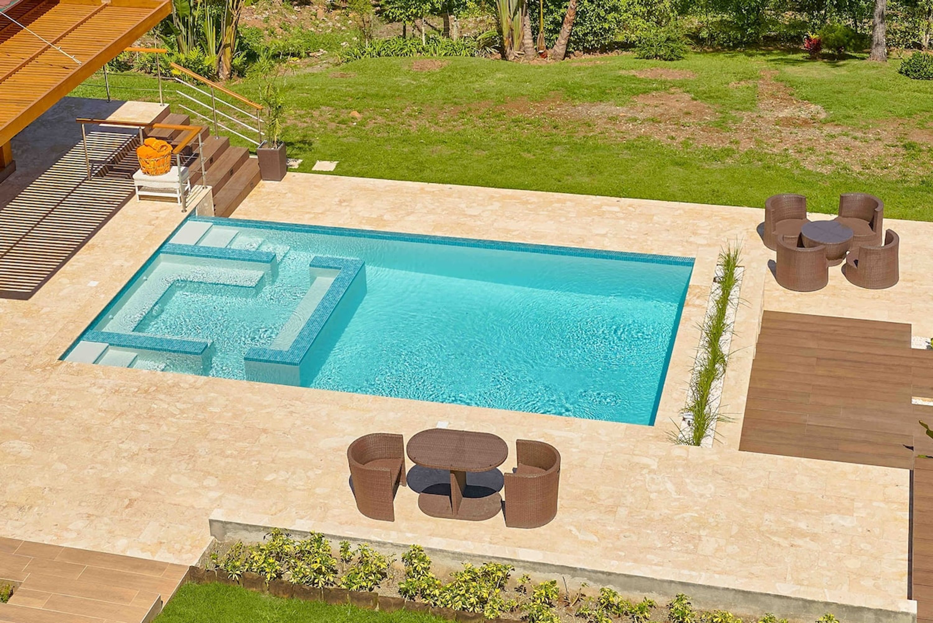 Une piscine rectangulaire ornée d’un motif géométrique, entourée d’une terrasse en pierre. On y trouve des meubles d’extérieur en osier et un petit espace abrité avec une table et des chaises.