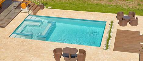 Une piscine rectangulaire ornée d’un motif géométrique, entourée d’une terrasse en pierre. On y trouve des meubles d’extérieur en osier et un petit espace abrité avec une table et des chaises.