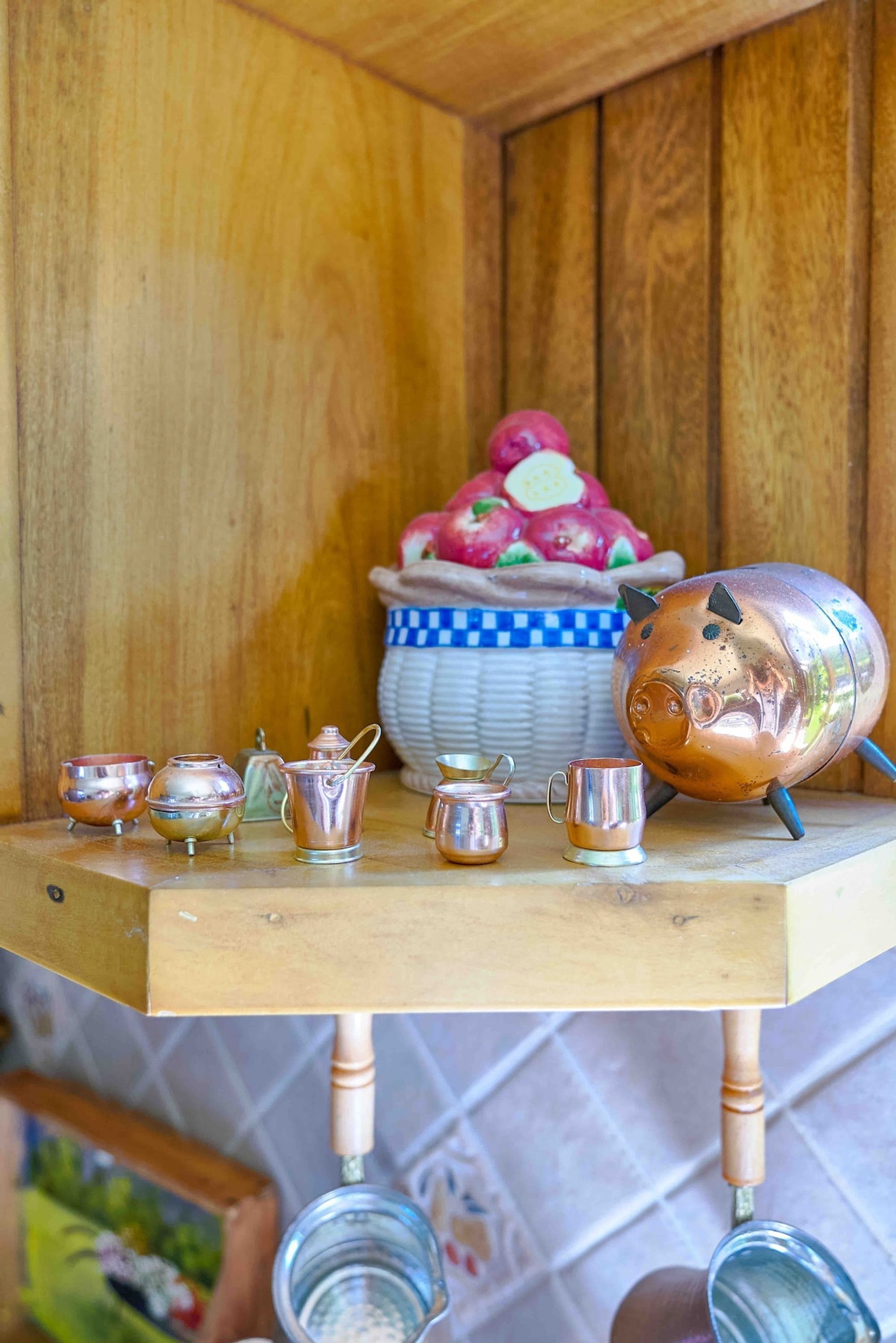 A wooden cabinet with a metal pig-shaped container on top, a basket of fruits, and several metal kitchen utensils on a wooden table.