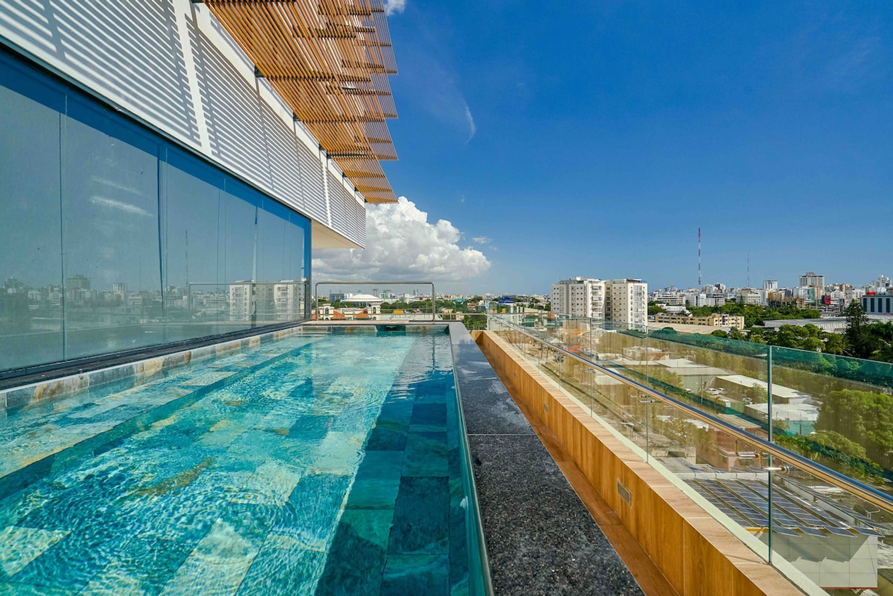 A rooftop infinity pool with a city view, a wooden pergola, and a glass railing.