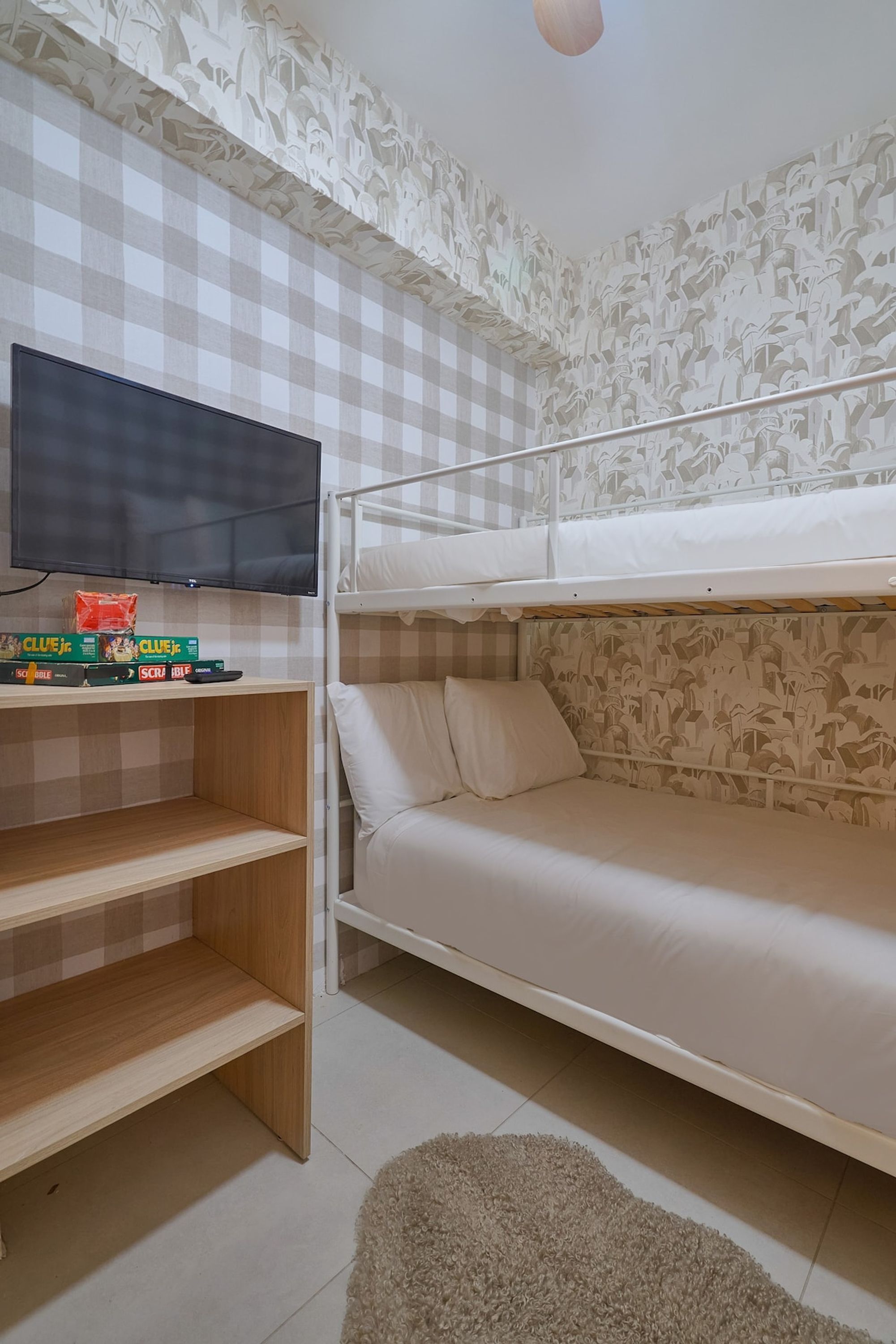 Una cama litera con un televisor encima, una estantería con libros y papel tapiz estampado.