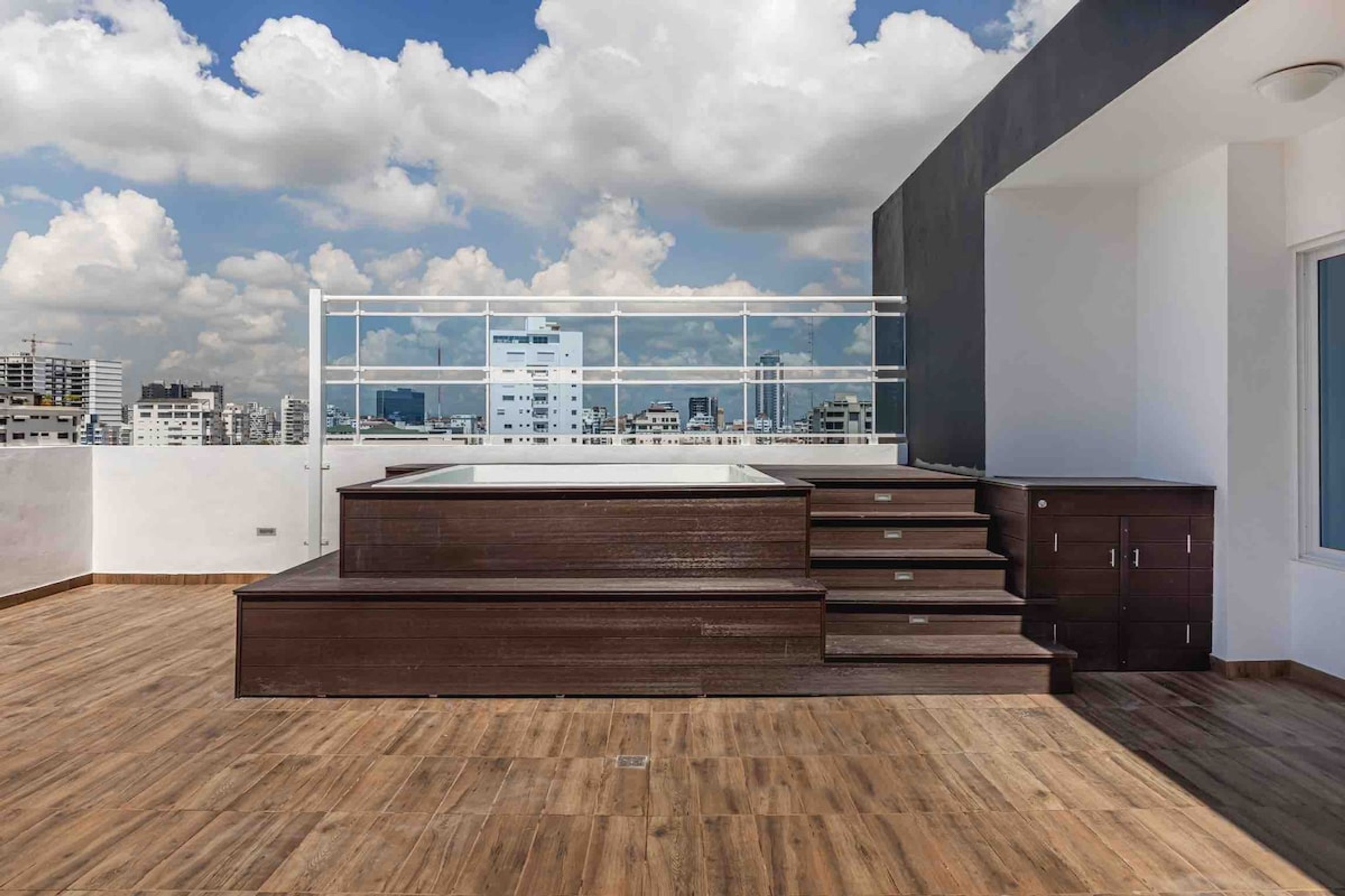 A modern rooftop terrace with a wooden floor, a glass railing, and a view of the city skyline.
