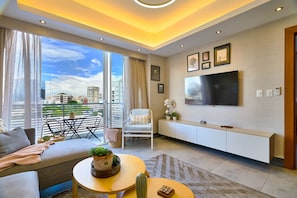 A modern living room with a sofa, a coffee table with a cactus, a TV mounted on the wall, and a balcony with a view of the city. - Piantini Views Downtown SD Gym Lounge Pool (Santo Domingo)