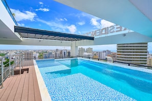 Une piscine à débordement située sur le toit, offrant une vue sur la ville, et entourée d’une terrasse en bois et de rampes en métal.