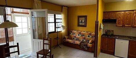 A compact kitchen with wooden cabinets, a dining table with chairs, a sofa, and a small refrigerator.