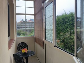 Uma varanda com mesa e cadeiras, vista para áreas verdes e edifícios, e uma janela com persiana texturizada.