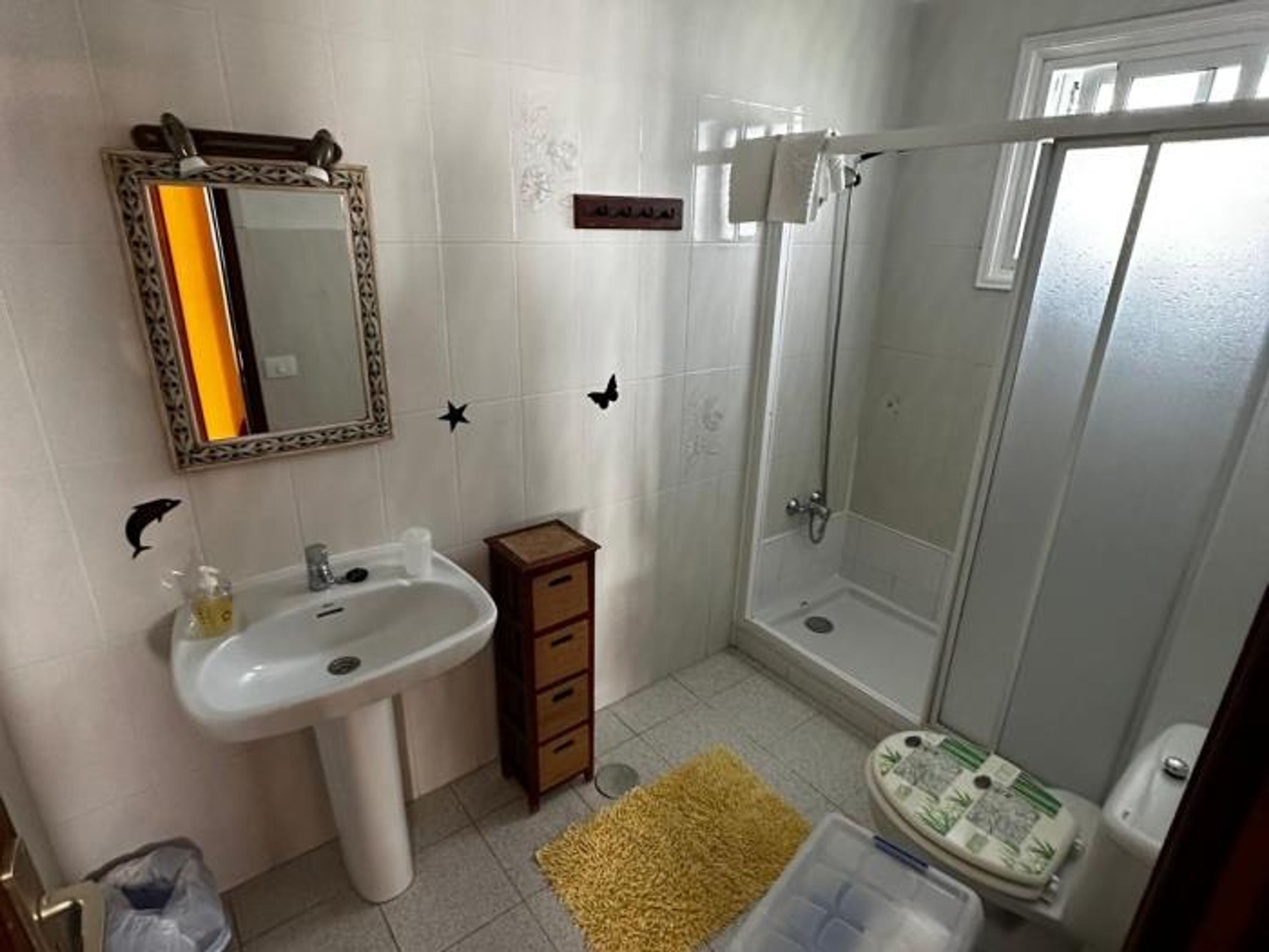 Une salle de bain avec un lavabo blanc, un miroir et une douche. On y trouve un petit meuble en bois et un tapis jaune sur le sol.