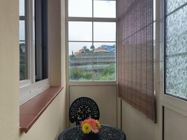 Una habitación con ventana que da a una zona residencial, una silla negra con respaldo decorativo y una mesita con un jarrón de flores.