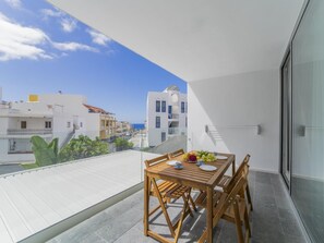 Un balcón con una mesa de madera dispuesta para dos personas, con vistas a una zona residencial con edificios blancos y un cielo azul despejado.