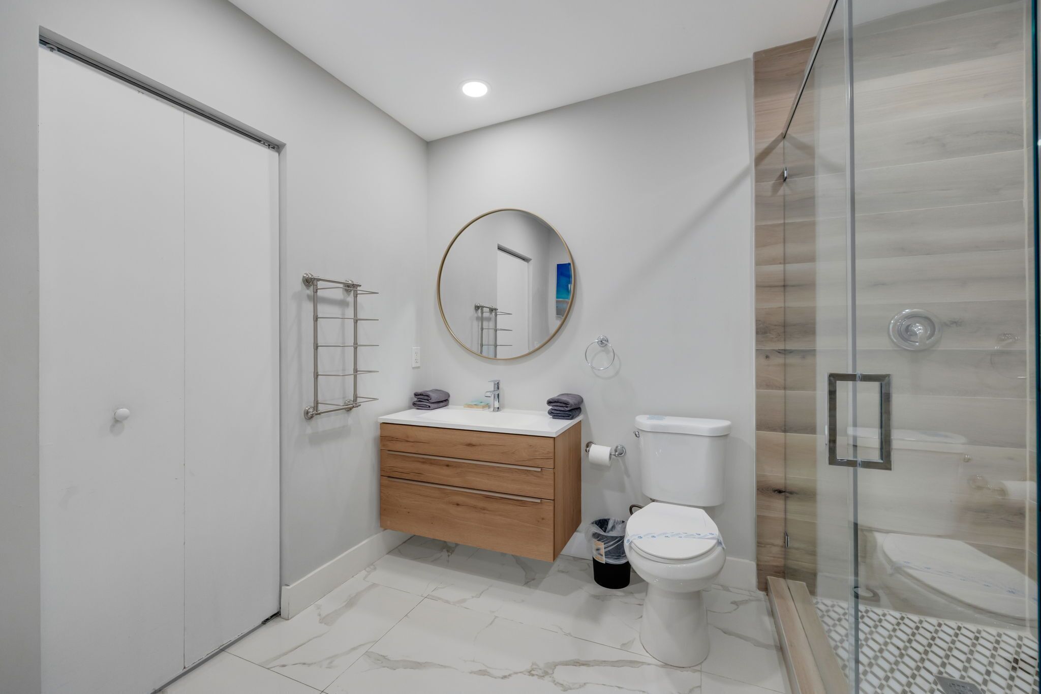 Et moderne badeværelse med et hvidt toilet, en trækommode, et rundt spejl og en brusekabine i glas.