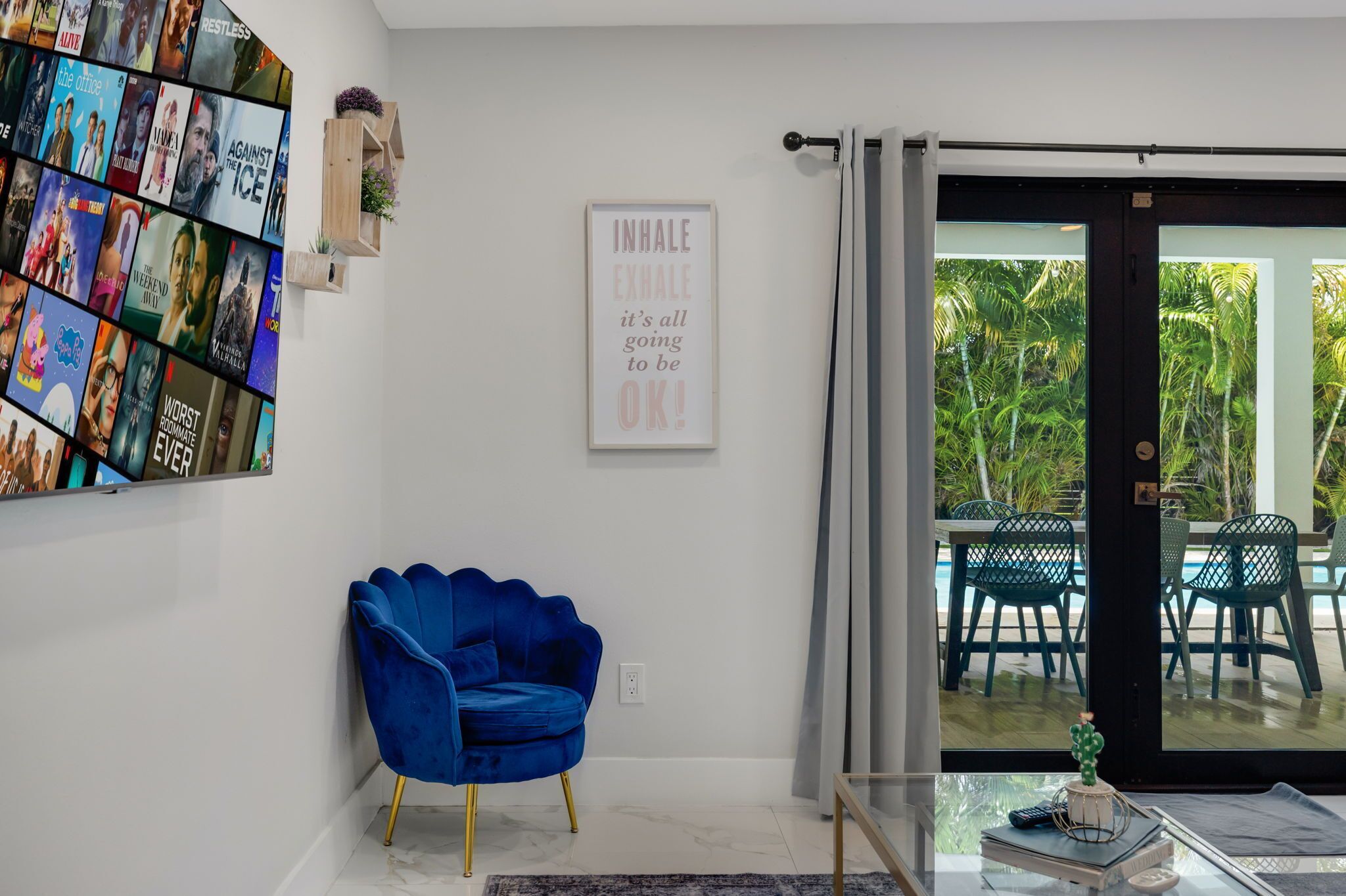 Een woonkamer met een blauwe stoel, een tv die aan de muur is bevestigd, en een schuifdeur die leidt naar een buitenruimte met stoelen.
