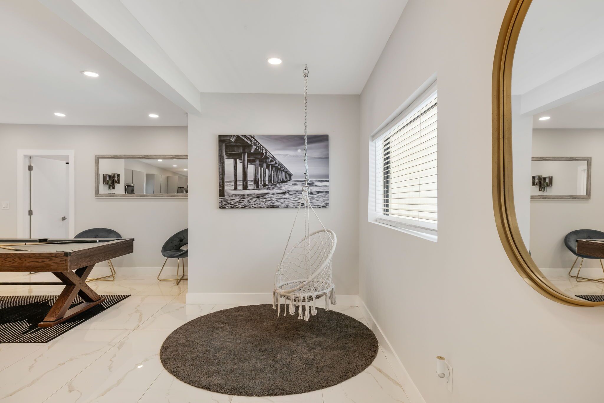 Um interior moderno com área de refeições, uma cadeira suspensa, um espelho grande e uma pintura de um cais.