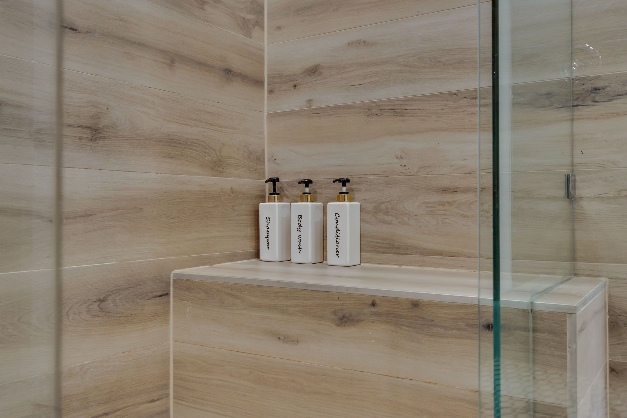 A bathroom with wooden panel walls, a glass shower enclosure, and a shelf with three toiletries.