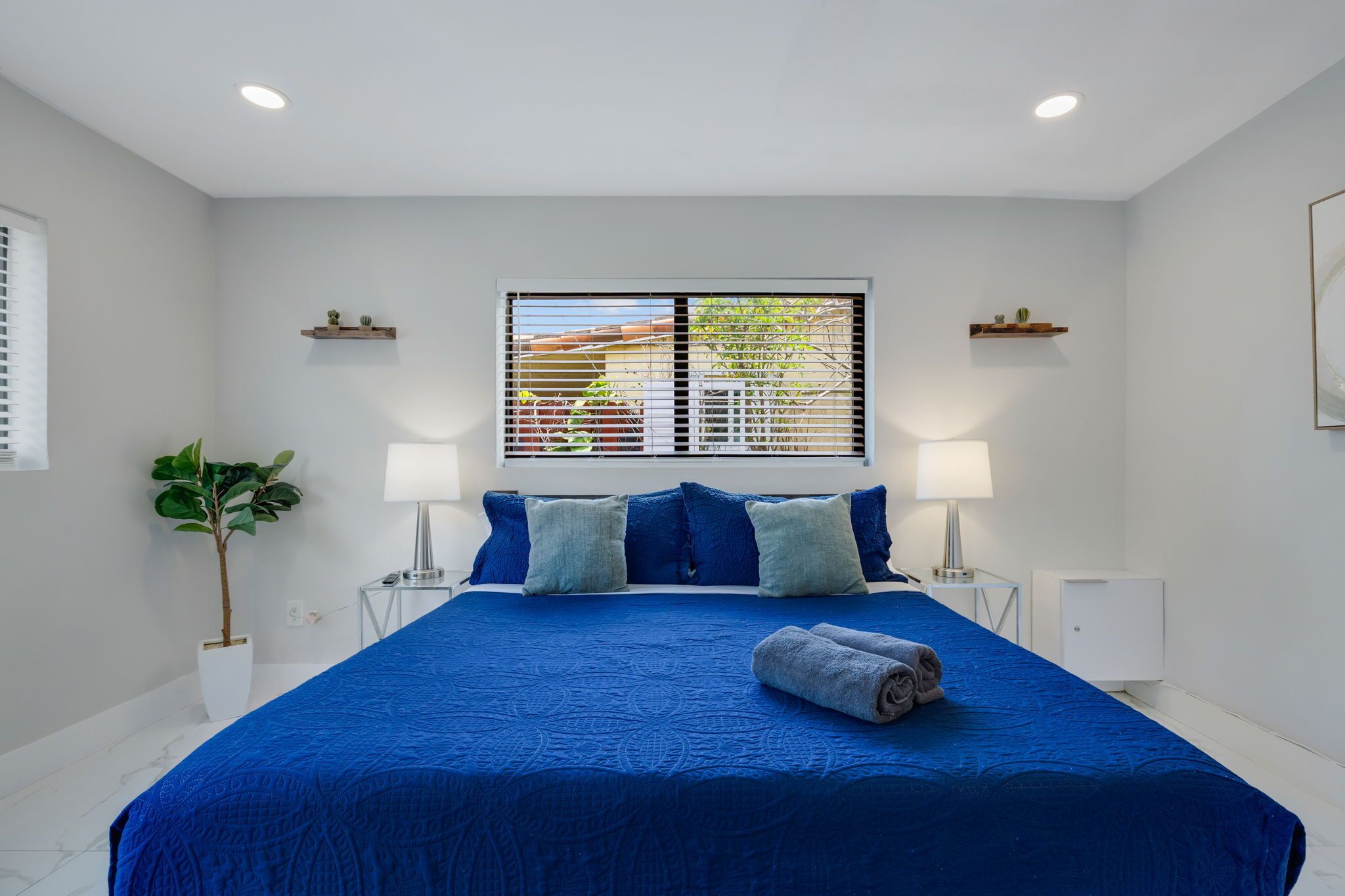 A bedroom with a large bed, two bedside tables, a window with blinds, a potted plant, and a wall-mounted shelf.
