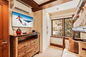 Une pièce avec une commode en bois, un téléviseur fixé au mur, une fenêtre donnant sur des arbres couverts de neige et un lit avec des oreillers.