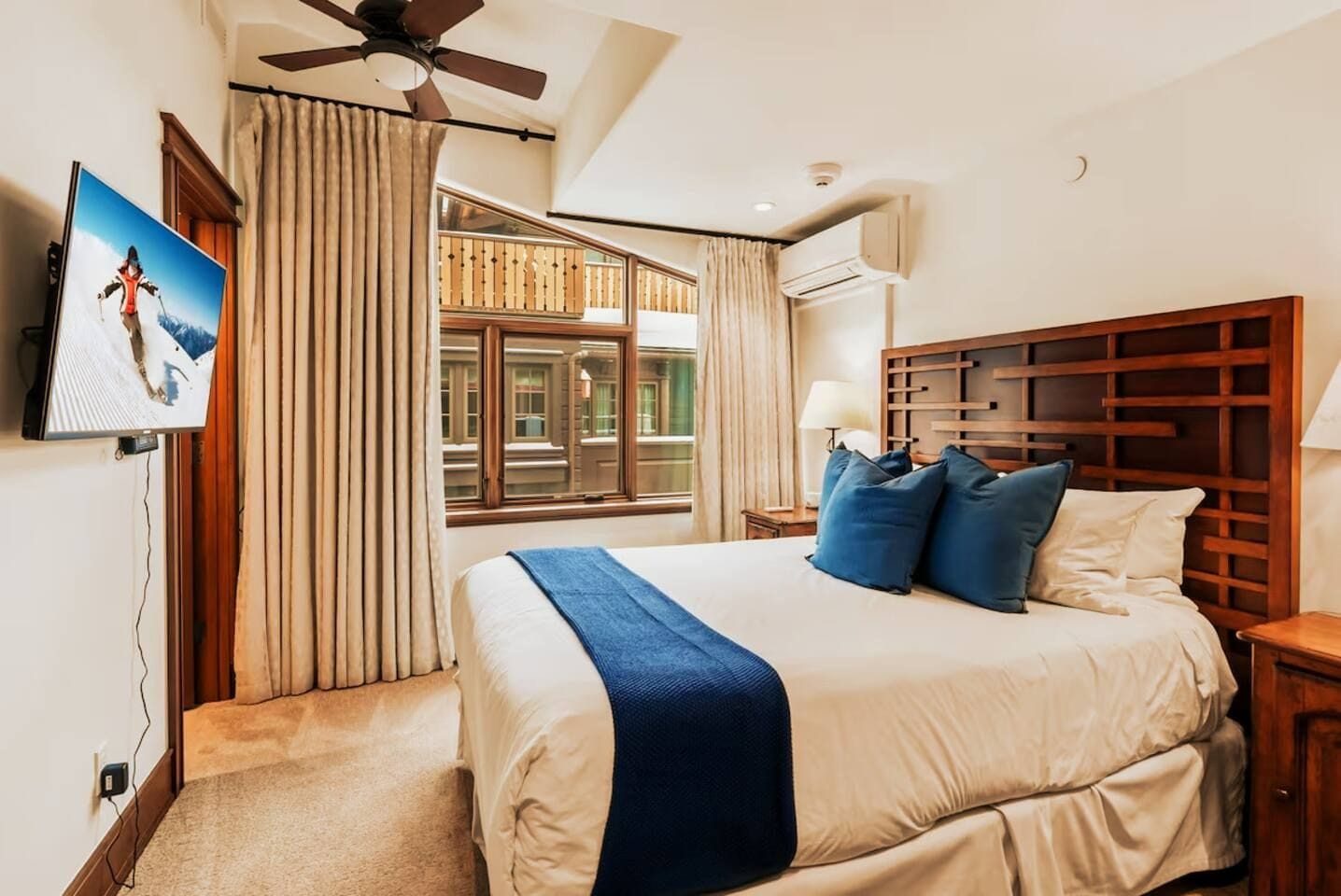 A hotel room with a bed, a television, a ceiling fan, a window with curtains, and an air conditioning unit.