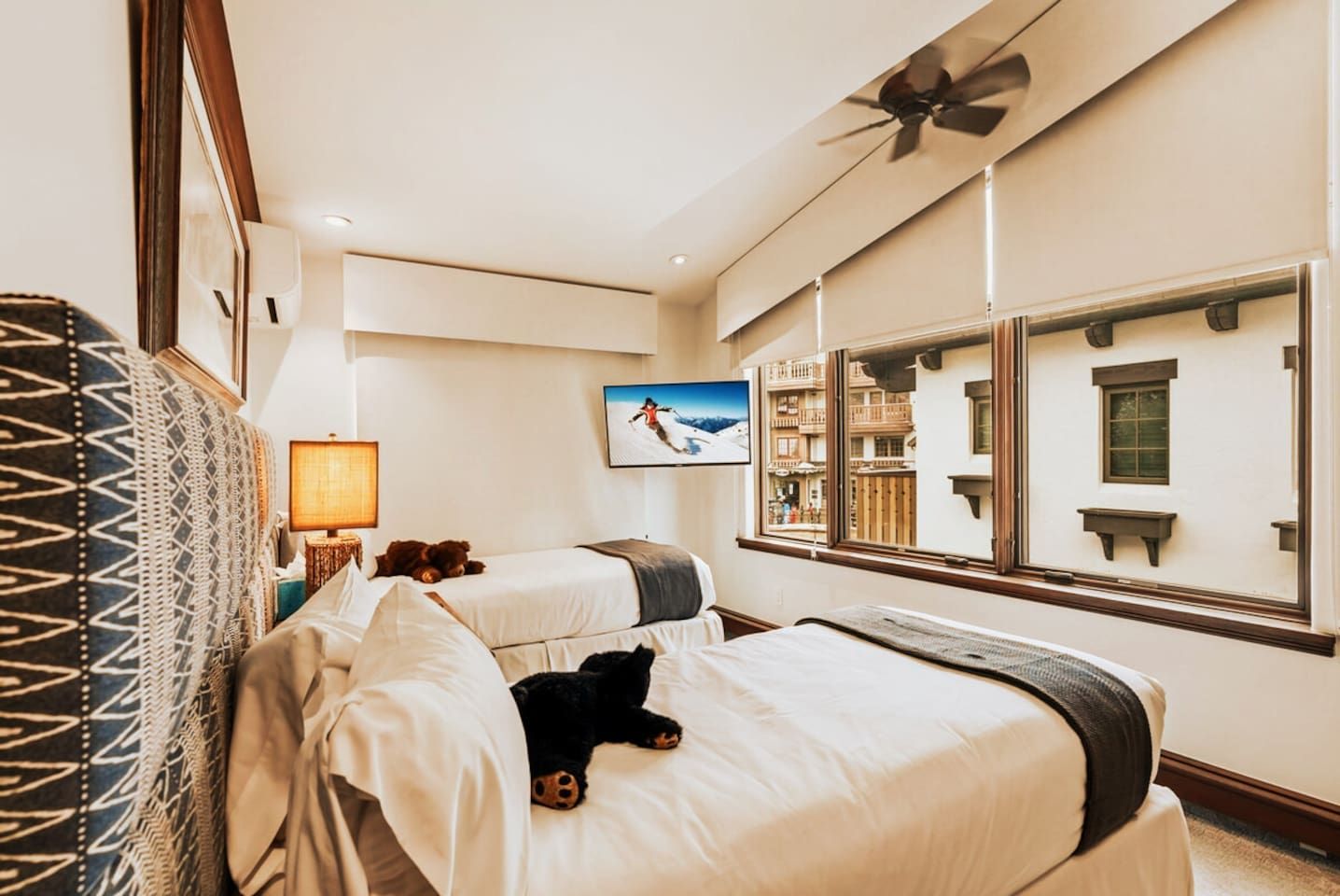 Ein Hotelzimmer mit zwei Betten, einem großen Spiegel, einem Deckenventilator und einem Fenster mit Blick auf ein Gebäude.