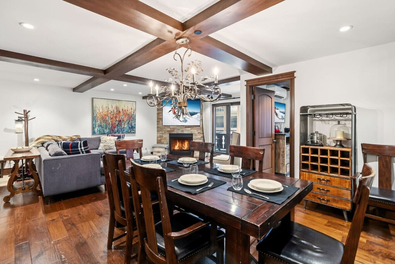 A dining area with a wooden table set for four, surrounded by wooden chairs. A fireplace is visible in the background, and a chandelier hangs from the ceiling.