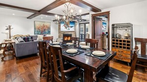 Un coin repas avec une table en bois dressée pour quatre personnes, entourée de chaises en bois. On aperçoit une cheminée au fond, et un lustre est suspendu au plafond.