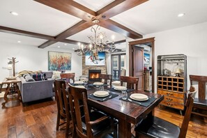 A dining area with a wooden table set for four, surrounded by wooden chairs. A fireplace is visible in the background, and a chandelier hangs from the ceiling. - Iconic Vail Village Retreat | Walk to Everything! (Vail)