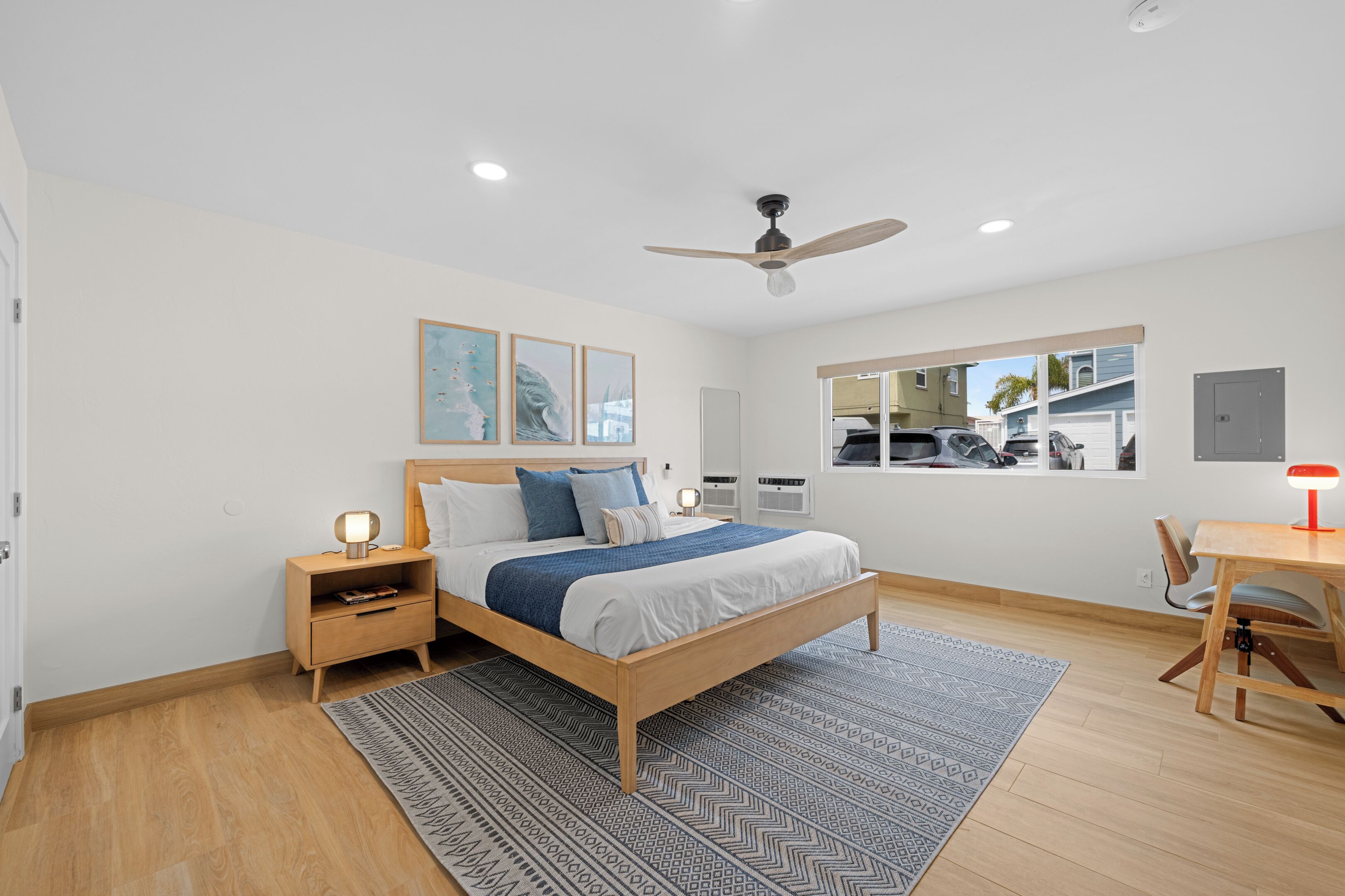 Un dormitorio con cama, mesitas de noche, un escritorio, una silla, ventilador de techo, ventana y obras de arte en la pared.
