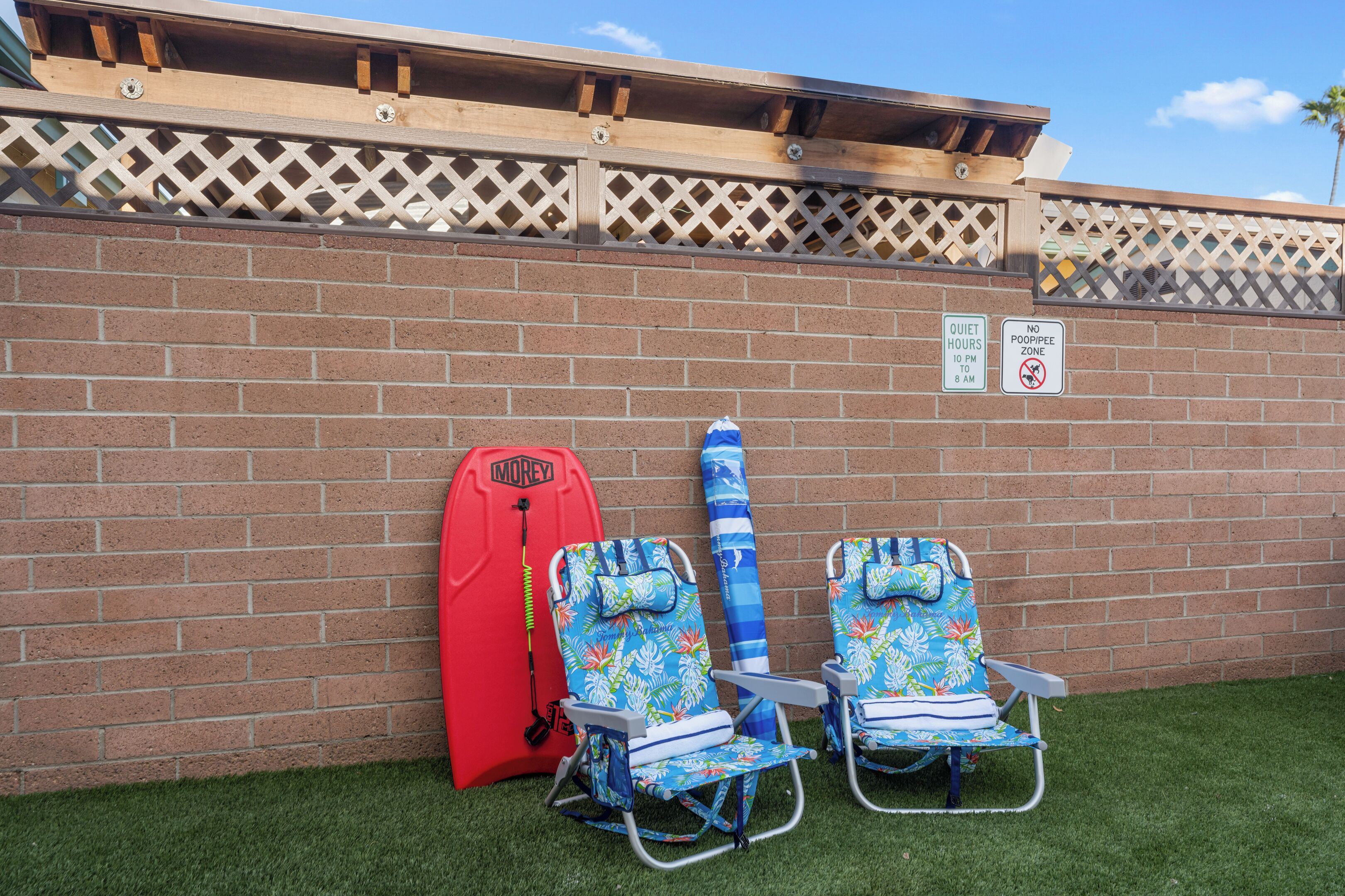 A wall with a brick pattern, two beach chairs, a red surfboard, and two signs indicating quiet hours and no poop/pee zone.