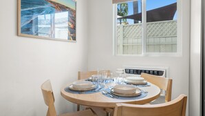 Ein Esstisch ist gedeckt mit Tellern, Gläsern und Besteck. An der Wand hängt ein Gemälde, und durch das Fenster ist ein Blick auf ein Gebäude und Palmen zu sehen.