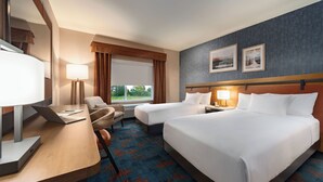 A hotel room with two beds, a desk with a laptop, a chair, a window with curtains, and framed pictures on the wall.