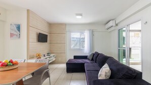 Un salon moderne avec un canapé foncé, une table à manger avec des chaises, un téléviseur fixé au mur et une grande fenêtre avec des rideaux.