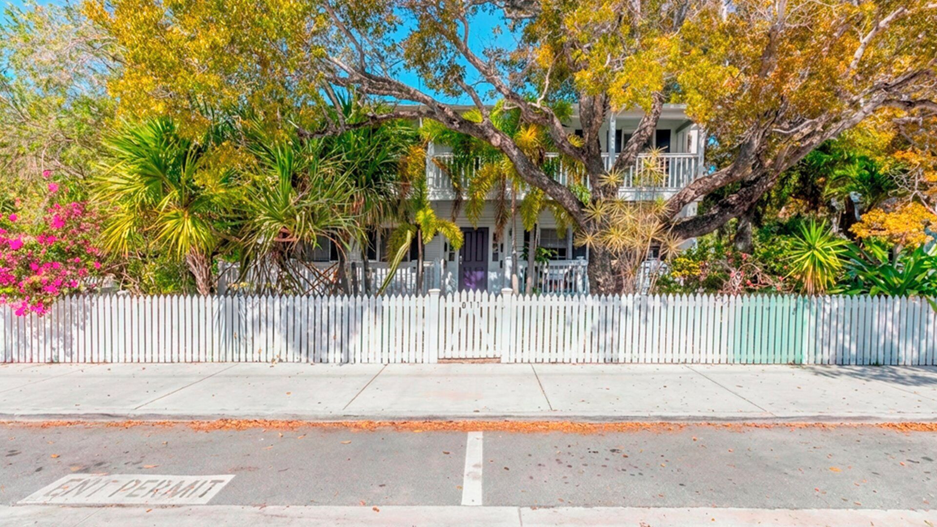 A two-story building with a white picket fence, surrounded by various tropical plants and trees.