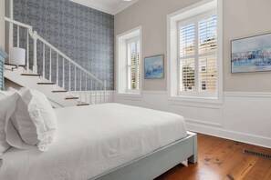 A bedroom with a large bed, a staircase, and two windows with blinds. - Best Location, 2bd in Heart of French Quarter! (Charleston)