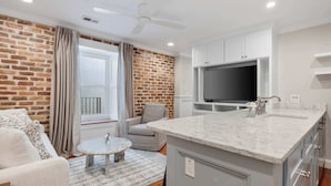 A modern kitchen with a marble countertop, white cabinets, and a flat-screen TV.