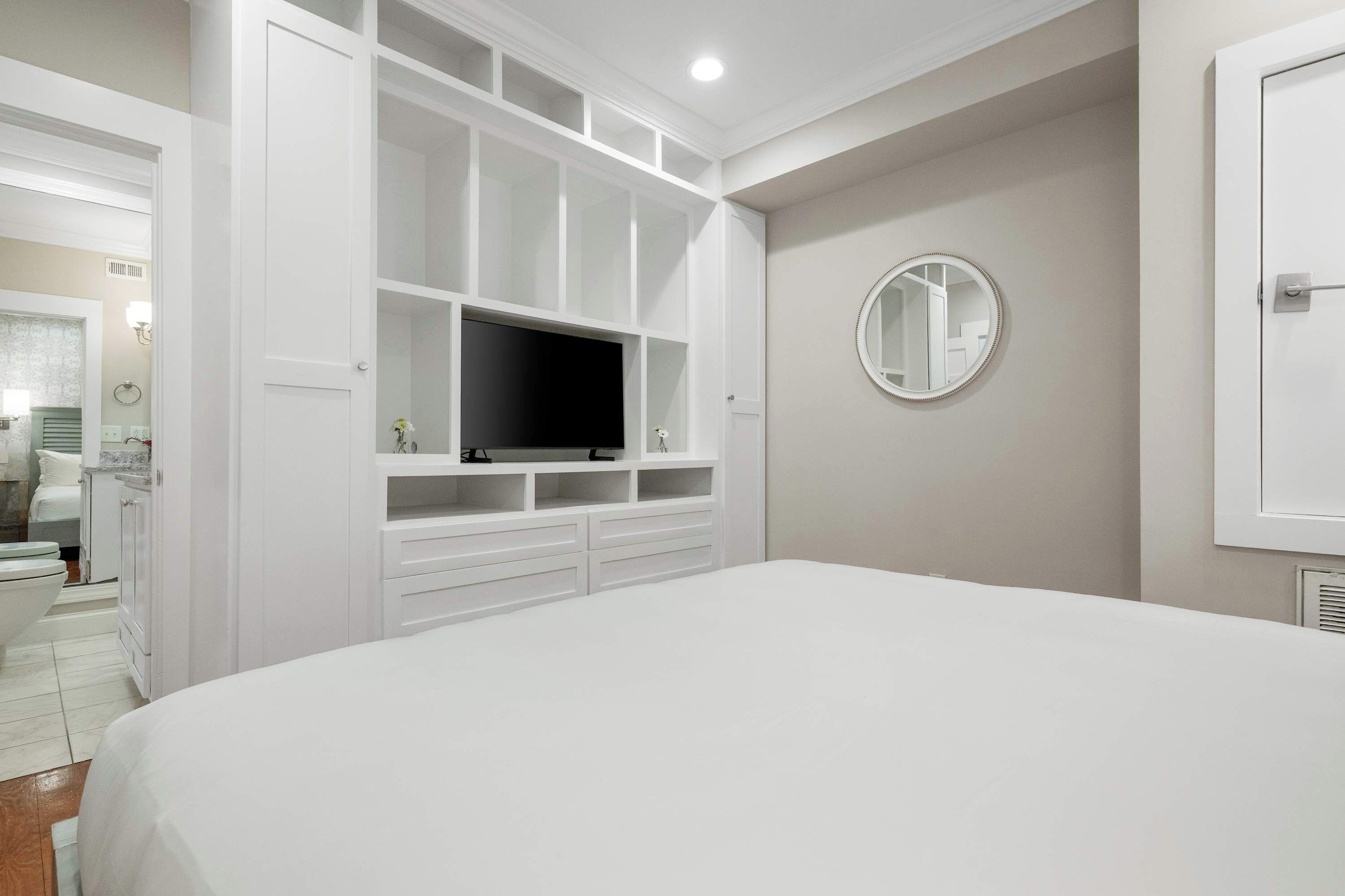 A bedroom with a large bed, built-in TV, storage cabinets, and a round mirror on the wall.