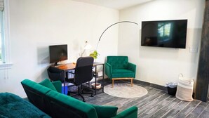 Moderni olohuone, jossa on työpöytä, kaksi vihreää nojatuolia, televisio ja suuri ikkuna.