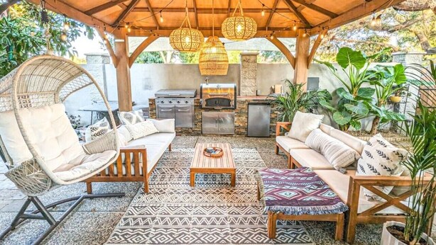 An outdoor patio with a wooden pergola, wicker furniture, a grill, and hanging lights.
