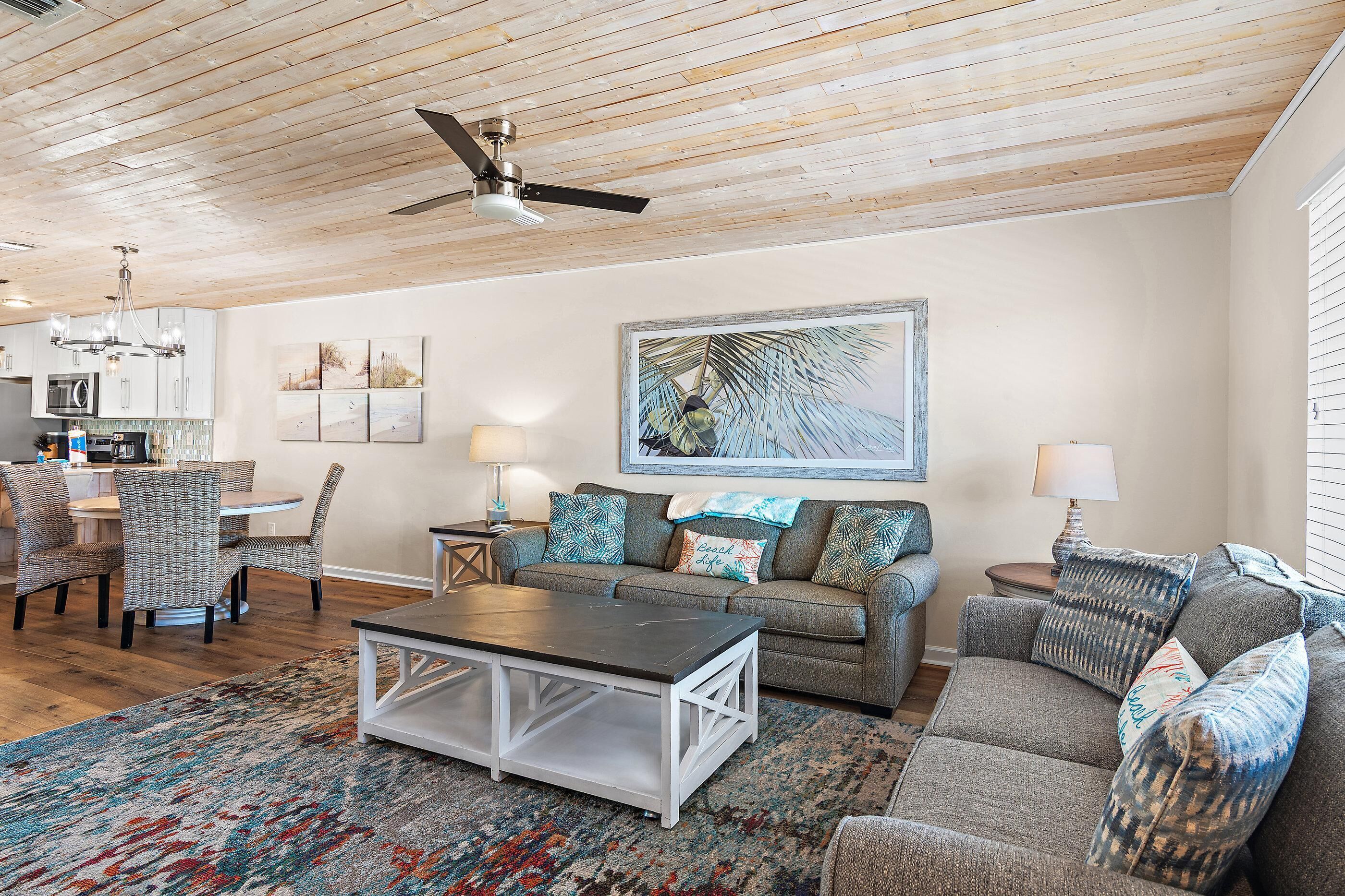 A living room with a ceiling fan, a dining area with a round table and chairs, and a kitchen in the background.