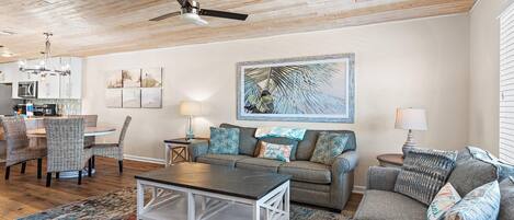 A living room with a ceiling fan, a dining area with a round table and chairs, and a kitchen in the background.