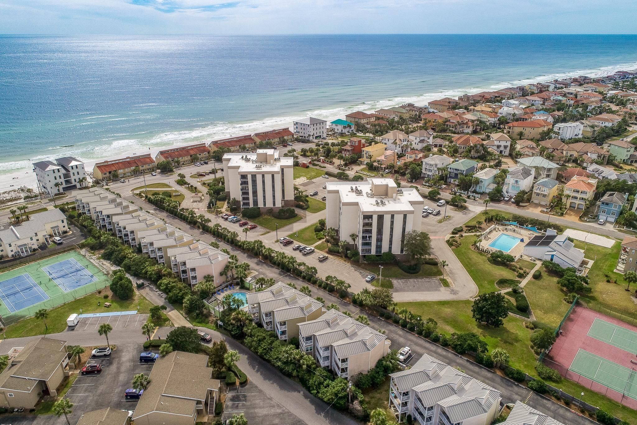 An aerial view of a coastal residential area with multiple buildings, a beach, and a tennis court.