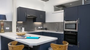 A modern kitchen with dark cabinets, a white countertop, a built-in oven, and two wicker chairs. - The Mayflower Suite (Plymouth)