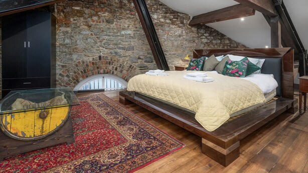 A bedroom with a stone wall, a bed with pillows, a nightstand, and a glass table with a rug. - The Mayflower Suite (Plymouth)