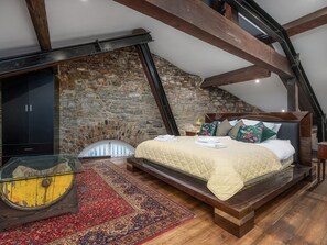 A bedroom with a stone wall, a bed with pillows, a nightstand, and a glass table with a rug.