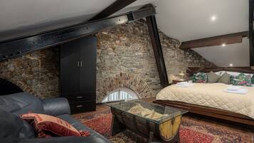 Dormitorio tipo loft con pared de piedra, cama con almohadones florales, sofá de cuero y mesa de centro de vidrio.