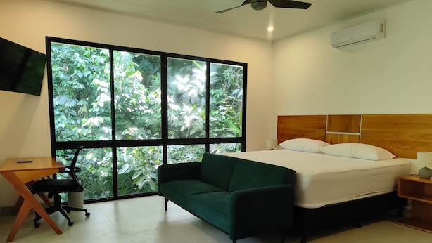 Een slaapkamer met een bed, een groene bank, een bureau en een groot raam met uitzicht op groen.