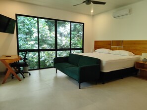 Un dormitorio con cama, sofá verde, escritorio y ventana grande con vista a vegetación.